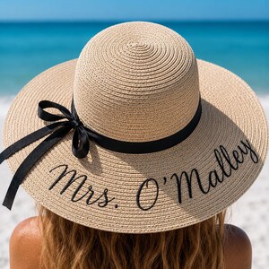 May include: A wide-brimmed, beige straw hat with a black ribbon and the embroidered text "Mrs. O'Malley". The hat is worn by a person with long, wavy brown hair, with a beach and ocean background.