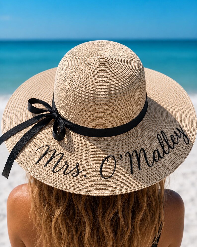 May include: A wide-brimmed, beige straw hat with a black ribbon and the embroidered text "Mrs. O'Malley". The hat is worn by a person with long, wavy brown hair, with a beach and ocean background.