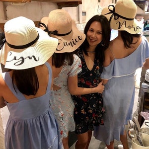May include: A group of people wearing straw sun hats with black ribbons and personalized names in black script. The hats have wide brims and are cream-colored. The people are wearing dresses in various colors and patterns.