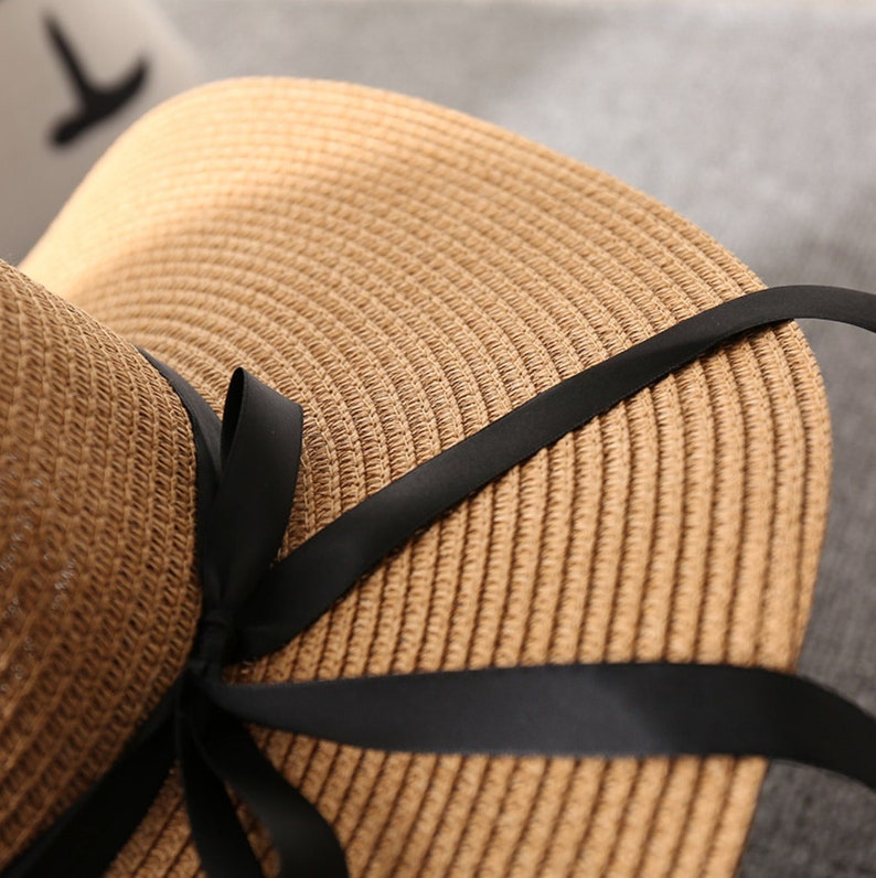 May include: A woven straw hat with a wide brim and a black ribbon tied in a bow. The hat is a light brown color and appears to be made of natural fibers. The black ribbon adds a contrasting accent to the hat.
