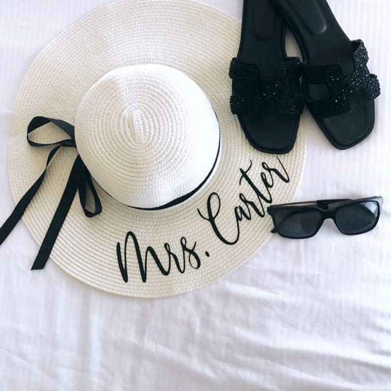 May include: A white sun hat with a black ribbon and the text 'Mrs. Carter' embroidered on the brim. The hat is lying on a white surface next to a pair of black sandals and black sunglasses.