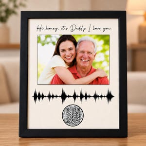 Puede incluir: Un marco de fotos negro que contiene una fotografía de una mujer y un hombre mayor. Sobre la foto está el texto "Hi honey, it's Daddy. I love you." Debajo de la foto hay un gráfico de onda sonora negro y un código QR circular en blanco y negro.