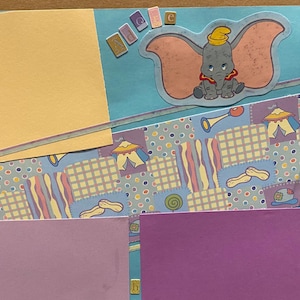 May include: A scrapbook page with a light blue background and a colourful patterned paper with a cartoon image of Dumbo the elephant. The page is decorated with yellow and purple ribbons and a blue and white striped ribbon.