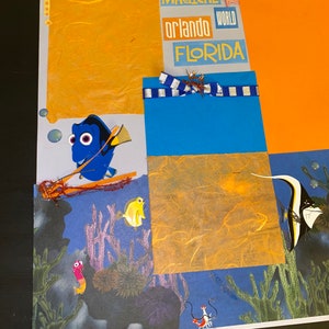May include: A scrapbook page with a blue and white striped ribbon, a yellow and orange paper background, and a blue fish with a red fin. The page also includes the text "MAGICAL", "ORLANDO WORLD", and "FLORIDA".
