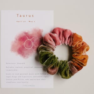 May include: A multi-colored velvet scrunchie in shades of pink, green, and orange. The scrunchie is next to a white card with the word "Taurus" and other text about the zodiac sign.