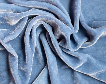 Op de afbeelding: Een close-up van een zachte, blauwe fluwelen stof met een gladde, zijdeachtige textuur. De stof is gedrapeerd en gevouwen, wat een luxe en elegante uitstraling creëert.