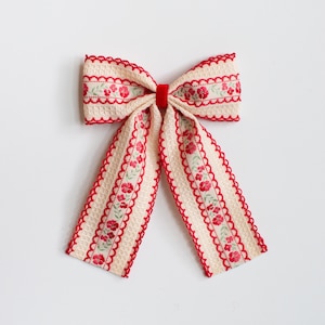 May include: A cream-colored bow with red scalloped edges and a red velvet center. The bow features a floral pattern with red flowers and green leaves. The ribbon has a textured waffle weave.