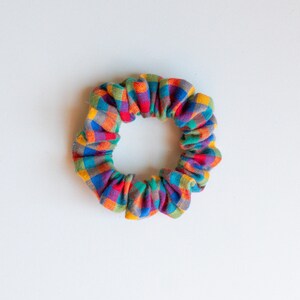 May include: A colorful scrunchie with a plaid pattern in shades of red, orange, yellow, blue, and green. The fabric is gathered and elasticized, creating a circular shape. This hair accessory is designed to hold hair in a ponytail or bun.
