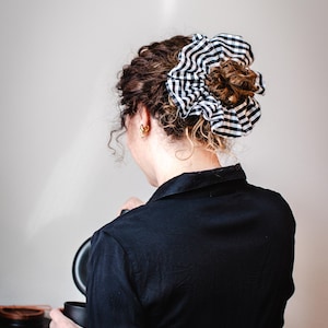 May include: A woman with curly brown hair, wearing a black shirt, and a black and white gingham scrunchie. The scrunchie is large and gathered, adding a stylish touch to the hairstyle. Gold hoop earrings are visible.