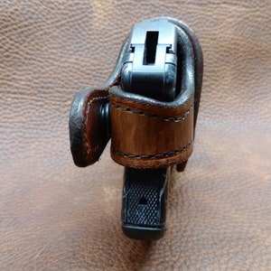 K&ouml;nnte beinhalten: Ein braunes Lederholster mit einer schwarzen Pistole darin. Das Holster hat eine G&uuml;rtelschlaufe und einen Druckknopfverschluss.