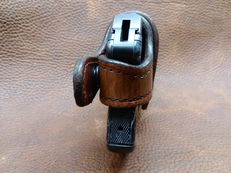 K&ouml;nnte beinhalten: Ein braunes Lederholster mit einer schwarzen Pistole darin. Das Holster hat eine G&uuml;rtelschlaufe und einen Druckknopfverschluss.