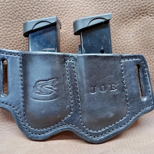 Może przedstawiać: Czarny skórzany uchwyt na magazynki z dwoma włożonymi magazynkami. Uchwyt posiada szytą konstrukcję, logo węża oraz wytłoczone z przodu imię "JOE". Zaprojektowany do przenoszenia dwóch magazynków.