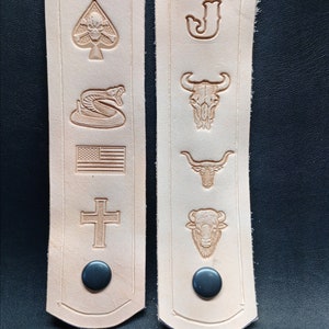 K&ouml;nnte beinhalten: Zwei hellbraune Lederriemen mit verschiedenen gepr&auml;gten Designs. Die Designs umfassen einen Totenkopf mit Knochen, eine Schlange, eine amerikanische Flagge, ein Kreuz, einen Buchstaben J, einen Stierkopf, einen Stier und einen B&uuml;ffel. Jeder Riemen hat einen schwarzen Knopf.