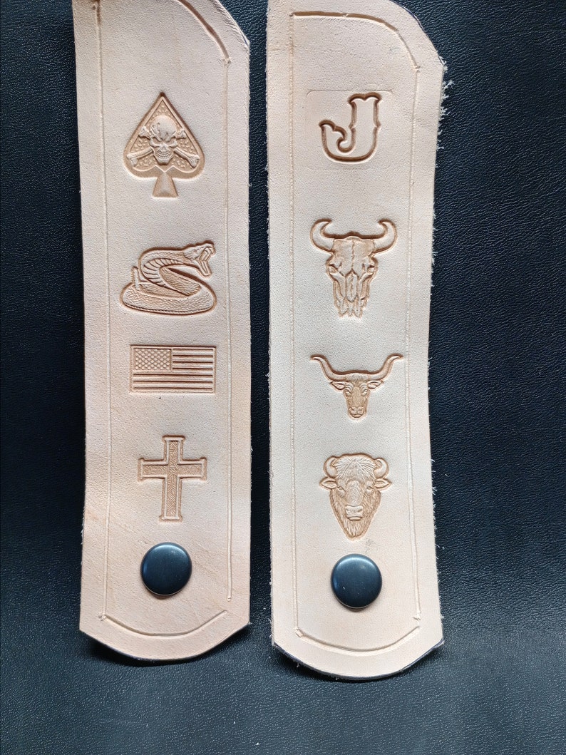 K&ouml;nnte beinhalten: Zwei hellbraune Lederriemen mit verschiedenen gepr&auml;gten Designs. Die Designs umfassen einen Totenkopf mit Knochen, eine Schlange, eine amerikanische Flagge, ein Kreuz, einen Buchstaben J, einen Stierkopf, einen Stier und einen B&uuml;ffel. Jeder Riemen hat einen schwarzen Knopf.