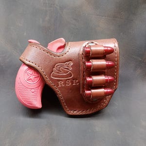 May include: A brown leather holster with a pink handgun and several red-tipped bullets. The holster features a snake emblem and the initials "RSR". The handgun has a star design on the grip.