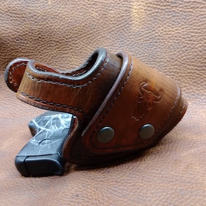 K&ouml;nnte beinhalten: Ein braunes Lederholster mit einer schwarzen Pistole darin. Das Holster hat ein gen&auml;htes Design und eine Metallschnalle. Das Holster liegt auf einer braunen Lederoberfl&auml;che.