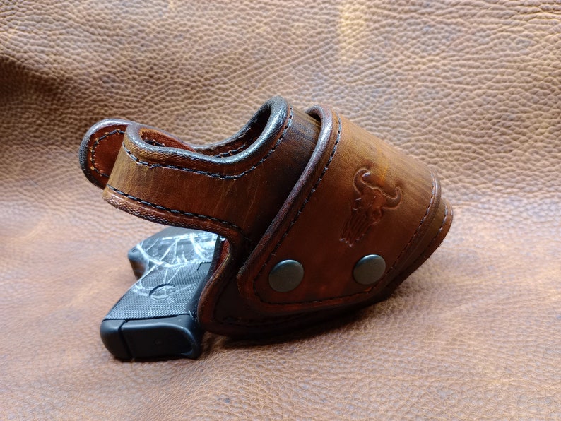 K&ouml;nnte beinhalten: Ein braunes Lederholster mit einer schwarzen Pistole darin. Das Holster hat ein gen&auml;htes Design und eine Metallschnalle. Das Holster liegt auf einer braunen Lederoberfl&auml;che.