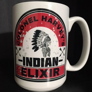May include: White ceramic mug with a red, black, and white logo. The logo features a Native American chief wearing a headdress and the text "Colonel Harvey's - Indian Elixir".