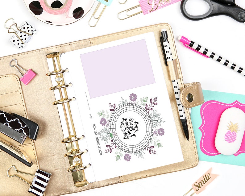 May include: A gold planner is open to a page with a lavender box and a circular floral design with the text "Look For The Good Every Day." Other stationery items include pens, paper clips, and a pink eraser with a pineapple design.