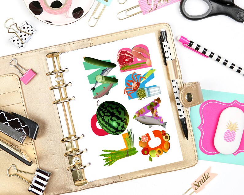 May include: A gold binder with a white page open to a page with the word "MEAL" spelled out in colorful food images. The page is surrounded by various office supplies, including a stapler, a pair of scissors, a pen, and a pink and black polka dot sticker.