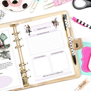 May include: A gold-colored planner with a white page open to a dream journal spread. The page features a whimsical illustration of Alice in Wonderland characters, including the Mad Hatter and the Cheshire Cat. The page is divided into sections for recording dreams, favorite dreams, and unpleasant dreams. The text "DREAM CONTROL" is printed at the top of the page.