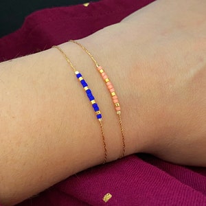 Peut inclure: Deux bracelets fins en chaîne dorée, chacun orné d'une rangée de petites perles colorées. L'un des bracelets présente des perles bleues, l'autre roses. Les bracelets sont présentés sur un poignet sur fond rose foncé.