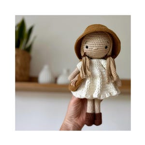 May include: A crocheted doll wearing a brown hat and a white dress. The doll has brown hair and brown eyes.