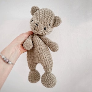 Könnte beinhalten: Ein handgefertigter, beigefarbener Häkelteddybär mit schwarzen Augen und Nase. Der Bär hat ein strukturiertes, plüschiges Aussehen mit abgerundeten Gliedmaßen und Ohren. Das Spielzeug wird vor einem weißen Hintergrund gehalten.