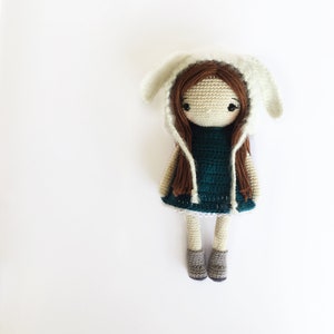 May include: A crocheted doll with brown hair wearing a white crocheted bunny hat and a teal dress. The doll is wearing gray shoes.