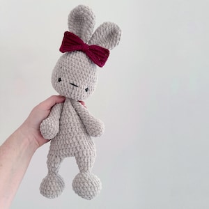May include: A light grey crocheted bunny toy with a dark red bow. The bunny has black stitched eyes and a closed mouth. The plush toy has long ears and rounded feet. The bunny is a soft, cuddly toy.