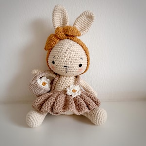 Puede incluir: Una muñeca de conejo de crochet que lleva un vestido marrón con flores blancas y una diadema marrón. El conejo lleva una pequeña bolsa de crochet con flores blancas.