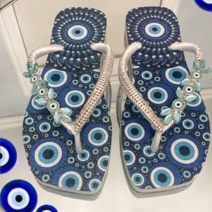 Peut inclure: Une paire de tongs bleues et blanches avec un motif d'yeux mal&eacute;fiques. Les tongs ont des lani&egrave;res argent&eacute;es et sont d&eacute;cor&eacute;es de perles bleues et blanches.