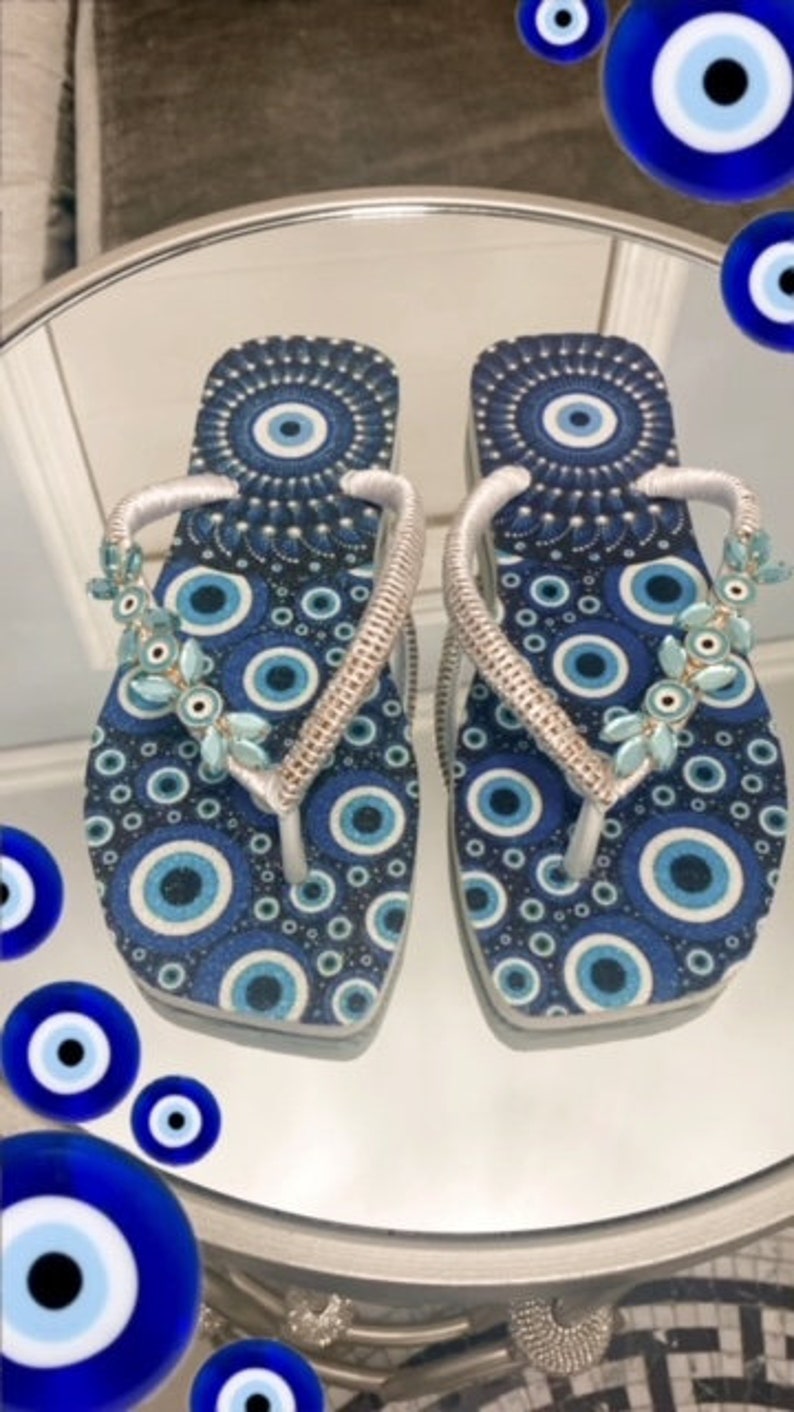 Peut inclure: Une paire de tongs bleues et blanches avec un motif d'yeux mal&eacute;fiques. Les tongs ont des lani&egrave;res argent&eacute;es et sont d&eacute;cor&eacute;es de perles bleues et blanches.