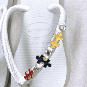 May include: White flip-flops with a white ribbon tied around the strap. The ribbon is decorated with colorful puzzle pieces and rhinestones.