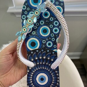 Peut inclure: Des tongs bleues et blanches avec une sangle blanche et un motif d'&oelig;il mal&eacute;fique bleu. La sangle est d&eacute;cor&eacute;e de strass bleus et dor&eacute;s.