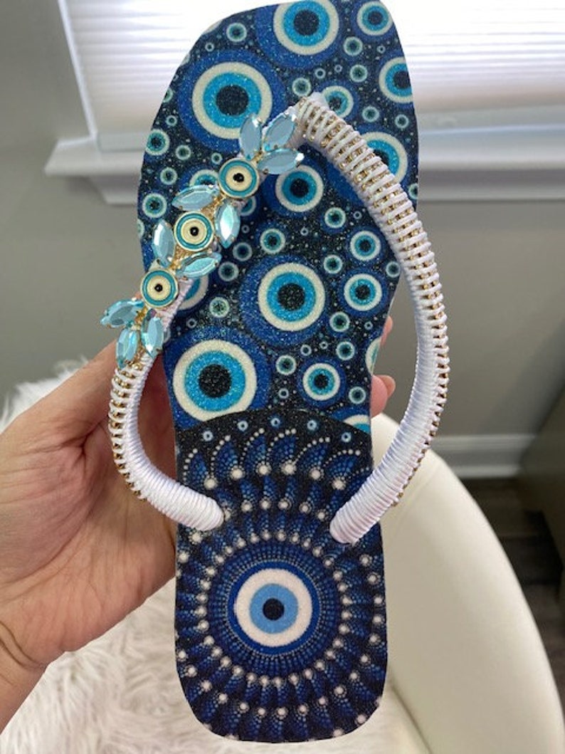 Peut inclure: Des tongs bleues et blanches avec une sangle blanche et un motif d'&oelig;il mal&eacute;fique bleu. La sangle est d&eacute;cor&eacute;e de strass bleus et dor&eacute;s.