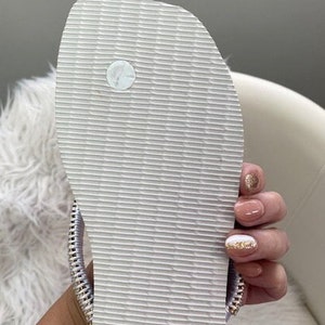 Peut inclure: Une tong blanche avec une semelle textur&eacute;e et une sangle argent&eacute;e. La tong a un petit cercle blanc sur le dessus de la semelle.