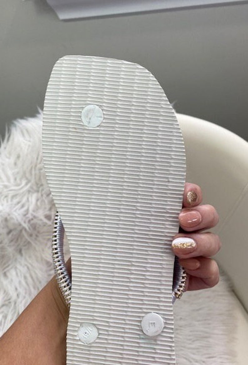 Peut inclure: Une tong blanche avec une semelle textur&eacute;e et une sangle argent&eacute;e. La tong a un petit cercle blanc sur le dessus de la semelle.