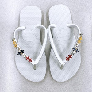 May include: White flip-flops with a white ribbon strap decorated with colorful puzzle pieces and rhinestones. The flip-flops have the brand name "Havaianas" printed on the straps.