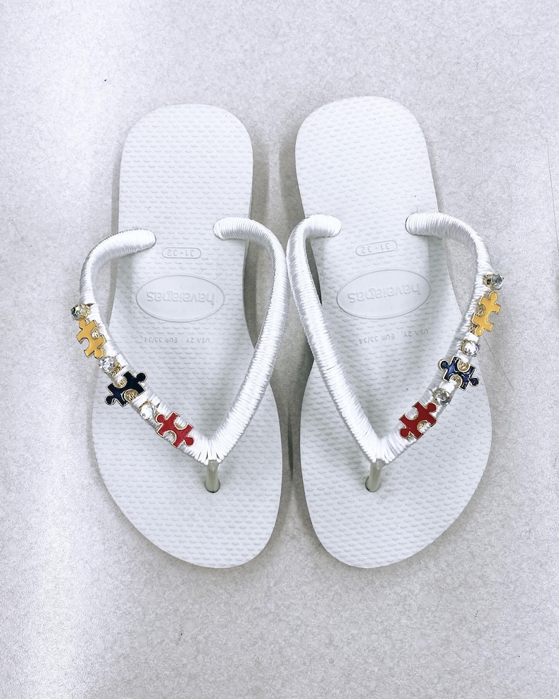 May include: White flip-flops with a white ribbon strap decorated with colorful puzzle pieces and rhinestones. The flip-flops have the brand name "Havaianas" printed on the straps.
