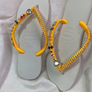 May include: A pair of white flip-flops with yellow straps decorated with colorful puzzle pieces and rhinestones. The straps are adorned with small, iridescent gems.