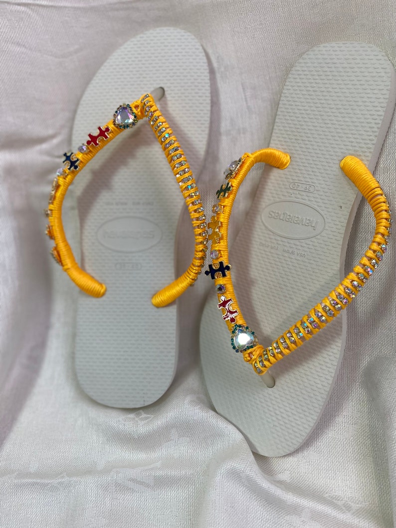 May include: A pair of white flip-flops with yellow straps decorated with colorful puzzle pieces and rhinestones. The straps are adorned with small, iridescent gems.