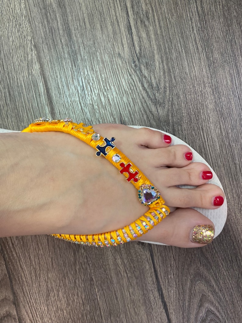 May include: Yellow and blue puzzle piece charm flip flops with rhinestones. The flip flops are white and have a gold heart charm with a purple rhinestone.