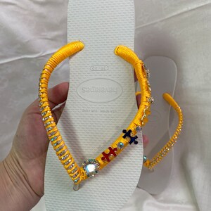 May include: White flip-flops with yellow straps decorated with rhinestones and colorful puzzle pieces. The straps are tied with a bow at the top. The flip-flops are labeled "Havaianas" and "Made in Brazil".