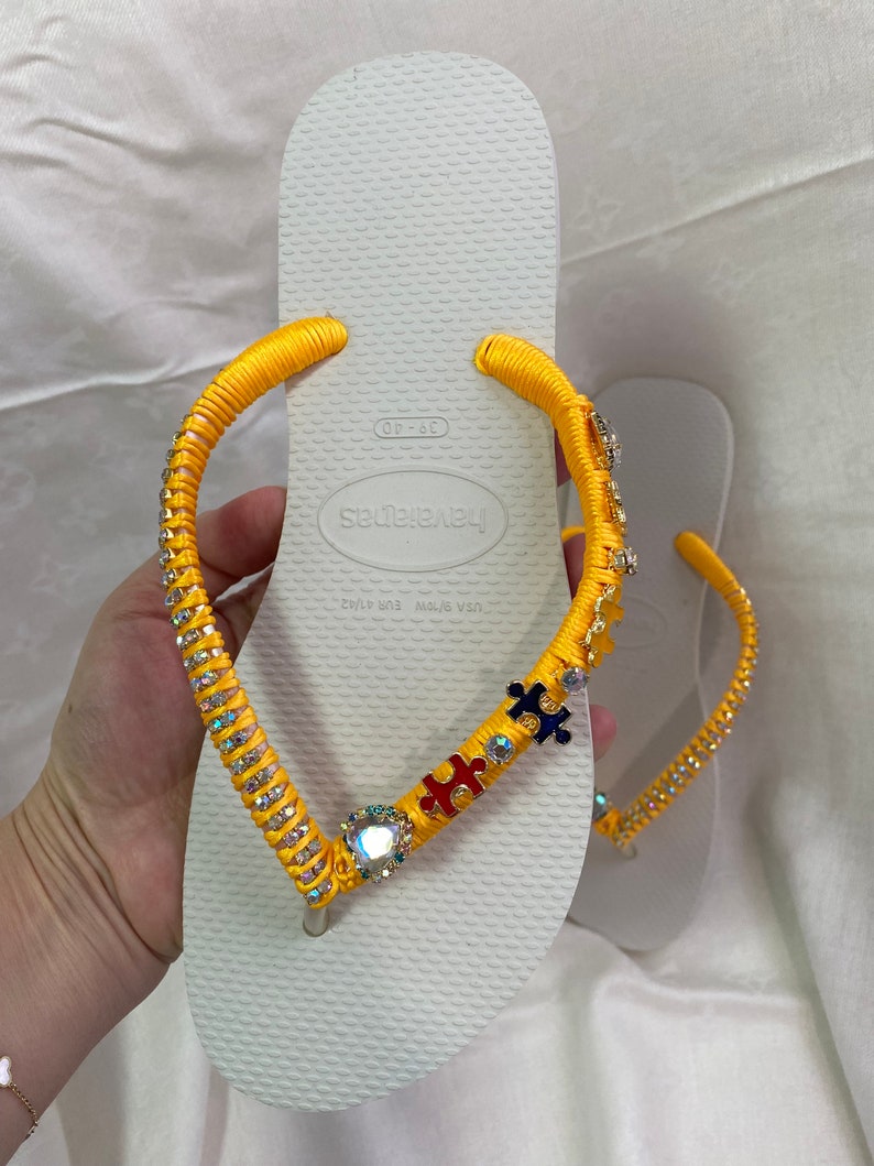 May include: White flip-flops with yellow straps decorated with rhinestones and colorful puzzle pieces. The straps are tied with a bow at the top. The flip-flops are labeled "Havaianas" and "Made in Brazil".