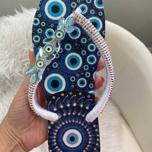 Peut inclure: Des tongs bleues et blanches avec une sangle blanche et un motif bleu de mauvais &oelig;il. La sangle est d&eacute;cor&eacute;e de perles bleues et dor&eacute;es.