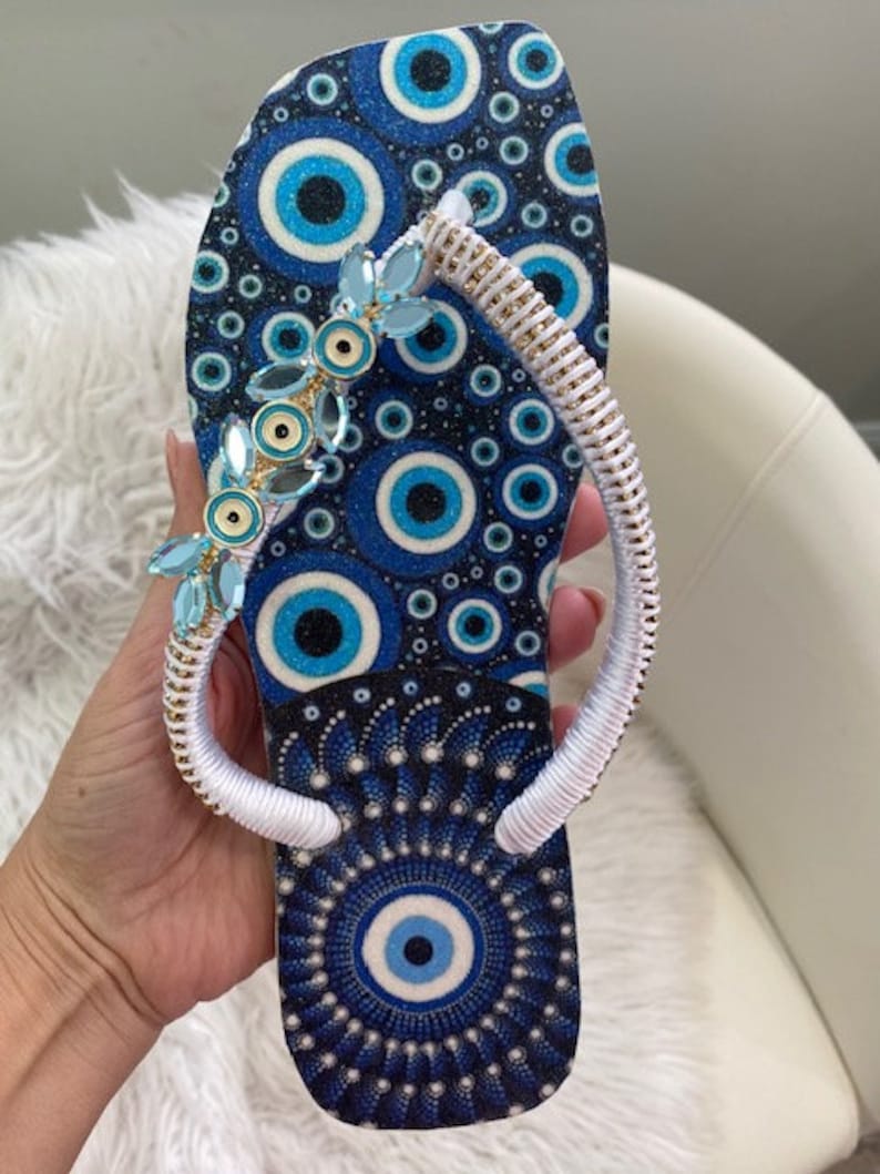 Peut inclure: Des tongs bleues et blanches avec une sangle blanche et un motif bleu de mauvais &oelig;il. La sangle est d&eacute;cor&eacute;e de perles bleues et dor&eacute;es.