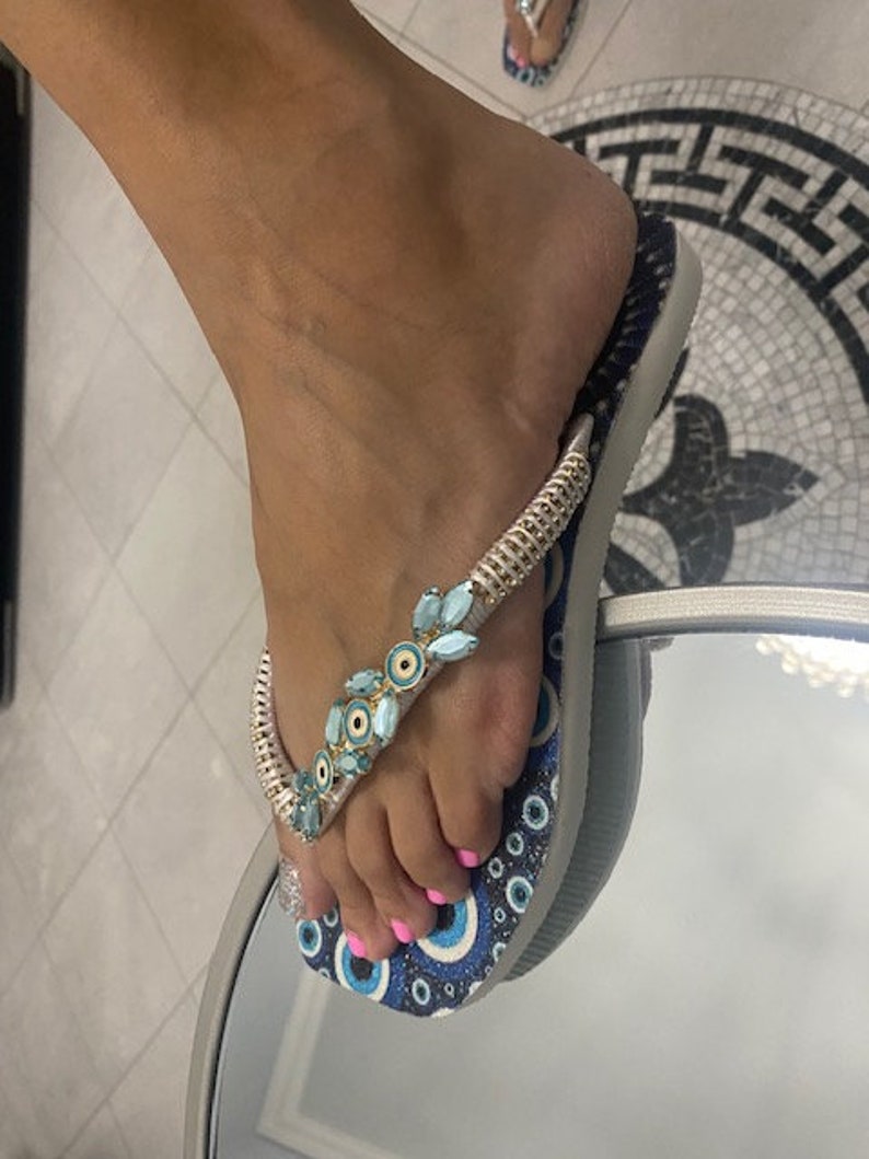Peut inclure: Des tongs bleues et blanches avec une cha&icirc;ne argent&eacute;e et des embellissements en forme d'&oelig;il bleu et blanc.