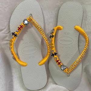 May include: A pair of white flip-flops with yellow straps decorated with rhinestones and puzzle piece charms. The flip-flops are size 39-40 and have the brand name "havaianas" printed on the sole.