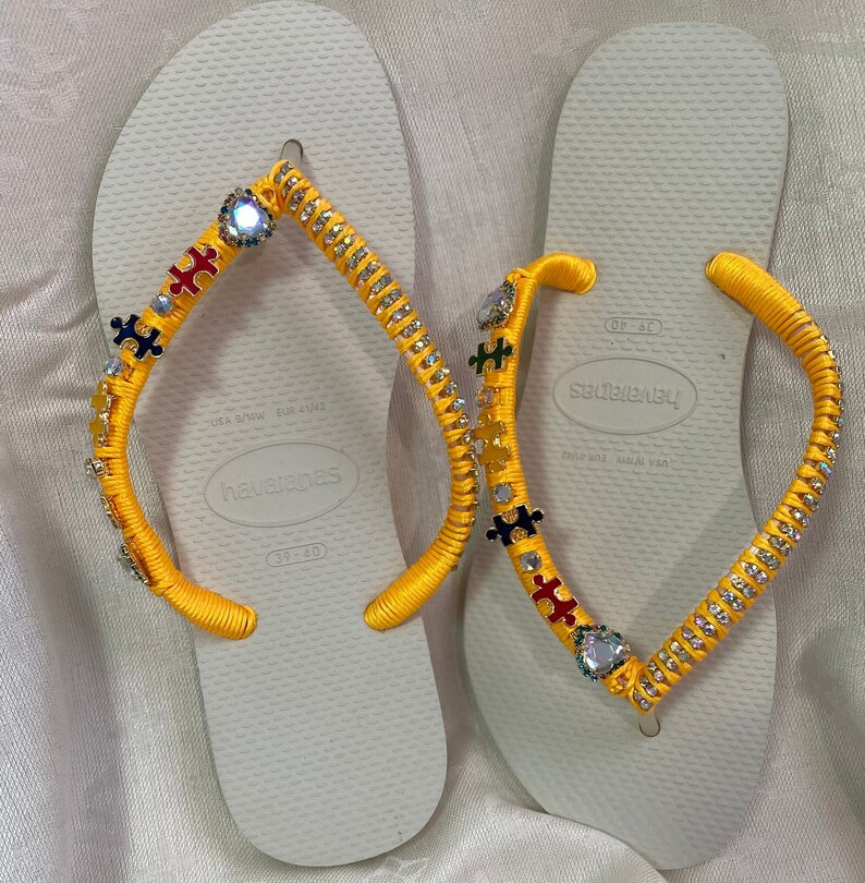 May include: A pair of white flip-flops with yellow straps decorated with rhinestones and puzzle piece charms. The flip-flops are size 39-40 and have the brand name "havaianas" printed on the sole.